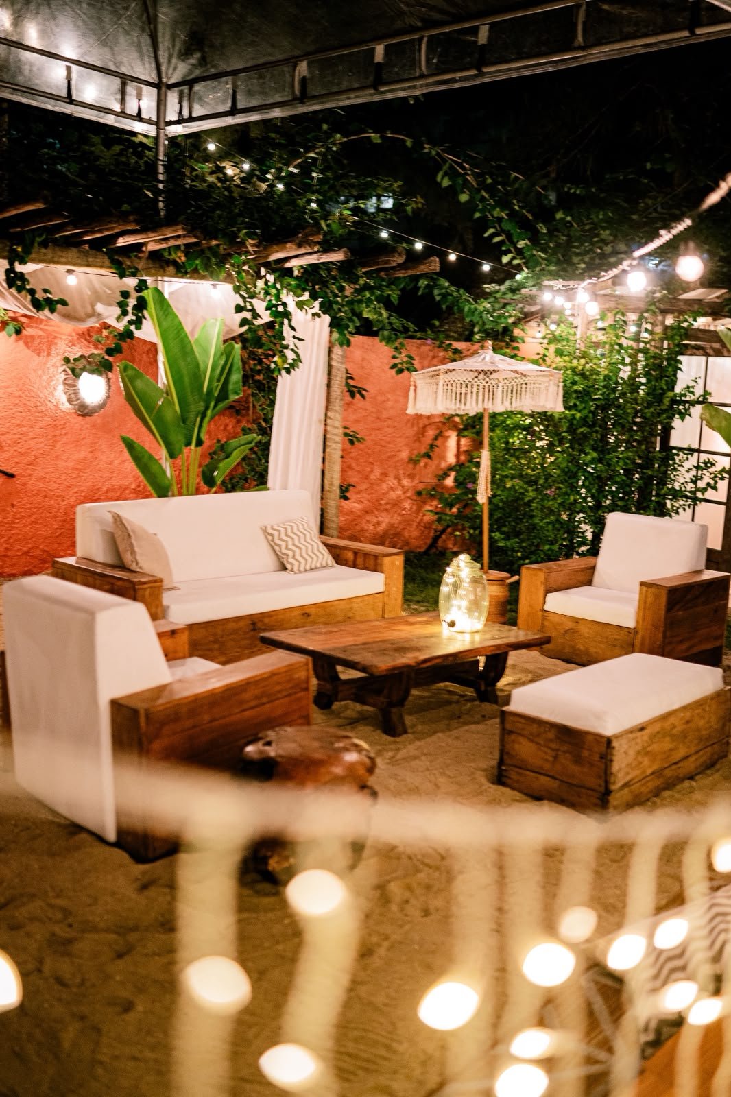 Lounge ao ar livre com sofás de madeira, almofadas brancas e luzes